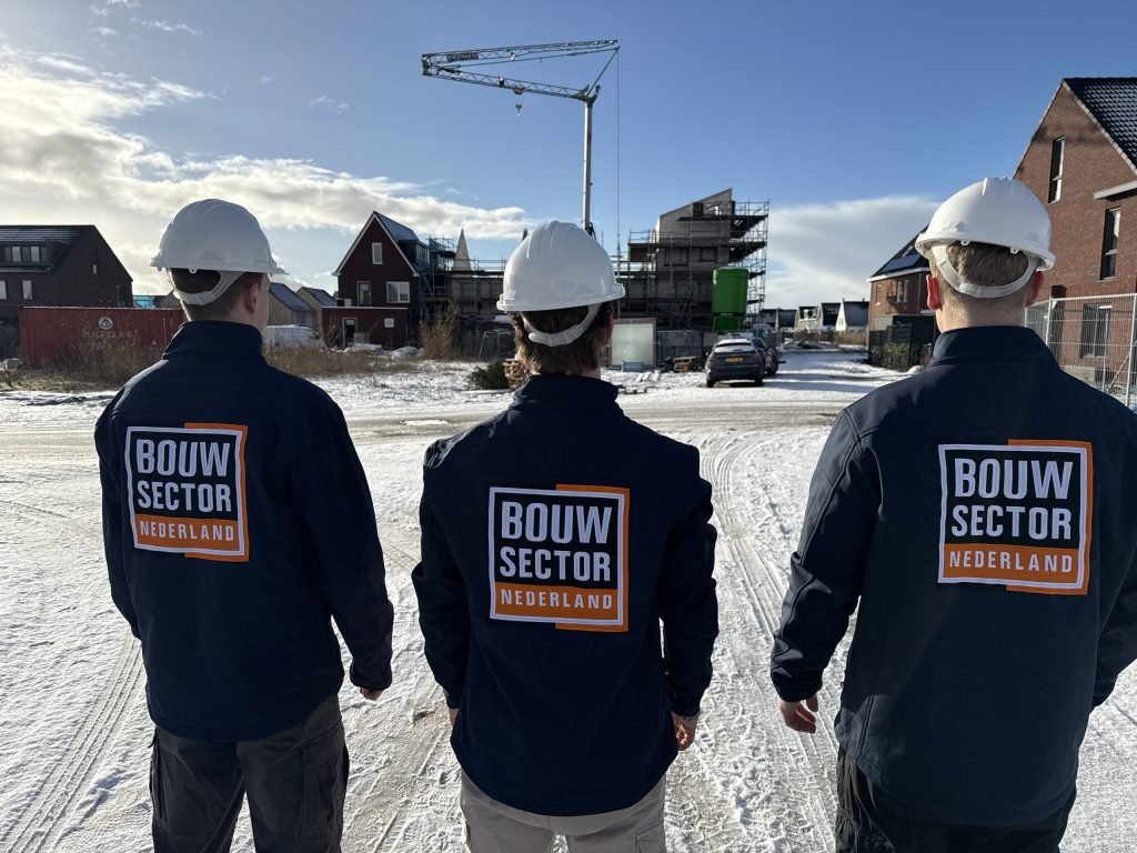 Drie vakmensen van Bouwsector Nederland met witte helm kijken uit over een straat met nieuwbouw in de sneeuw